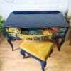 A dark blue vintage French style dressing table adorned with gold and multicolored floral patterns on the drawers stands against a white brick wall. One drawer is slightly open, and it is accompanied by a matching dark blue stool with yellow cushioning, all positioned on a wooden floor.