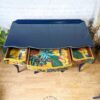 A vibrantly painted vintage French style dressing table features open drawers and displays an intricate design of tropical birds, plants, and fruits. The tabletop is dark blue with a scalloped edge. This beautiful piece stands on a hardwood floor against a white brick wall.