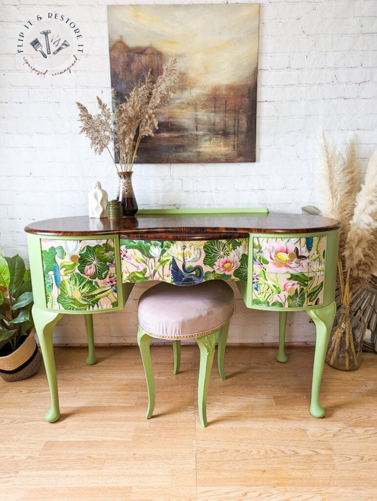 A Queen Anne painted walnut dressing table with a brown top and floral-patterned drawers stands against a brick wall. In front of the dressing table, there is a round, light pink upholstered stool from the matching set. Decorative vases with dried foliage adorn the tabletop, while wall art hangs above it.