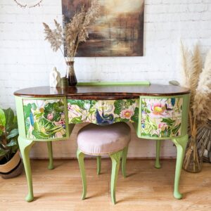 A Queen Anne painted walnut dressing table with a brown top and floral-patterned drawers stands against a brick wall. In front of the dressing table, there is a round, light pink upholstered stool from the matching set. Decorative vases with dried foliage adorn the tabletop, while wall art hangs above it.