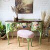 A chic vanity set against a white brick wall features the Queen Anne Painted Walnut Bedroom Set, which includes a green and floral-patterned dressing table/vanity along with a matching pink cushioned stool. Decorative items, including a plant and dried floral arrangements, complement the stylish and artistic ambiance.