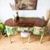 A Queen Anne Painted Walnut Dressing Table, featuring a polished dark wooden top and green legs, boasts three drawers decorated with vibrant floral and bird patterns. Atop the table sits a green vase alongside other small decor items. Nearby, a potted plant and dried floral arrangement add to the charming scene.