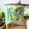 A vintage Queen Anne Painted Walnut Dressing Table/Vanity with a stool. The vanity has two drawers and features vibrant floral and leaf patterns on its green surface. The top is dark wood with a glossy finish. Surrounding the vanity are various plants, and in the background is a white brick wall. A painted logo reads "Flip It & Restore It.