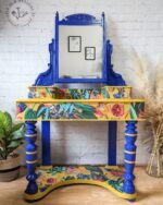 The Vintage Victorian Mahogany Duchess Dressing Table Painted Bedroom Set features a wooden vanity with a large mirror framed in an ornate blue design. The vanity top displays a vibrant floral and bird pattern. Positioned against a white brick wall, the set is flanked by lush plants on either side, while small artworks adorn the surrounding wall space.