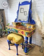 Introducing the Vintage Victorian Mahogany Duchess Dressing Table Painted Bedroom Set: A vibrantly decorated vanity set featuring a tall mirror with an intricate blue frame, a polished wooden tabletop adorned with vibrant floral patterns, and an upholstered stool in mustard-yellow velvet. Perfectly showcased against a white brick wall.