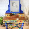 A vibrantly painted Vintage Victorian Mahogany Duchess Dressing Table with a large mirror. The mirror's frame and legs are primarily blue with intricate carvings, while the drawers and lower shelf feature bold floral designs in yellow, green, and red. Various plants and framed art are in the background.