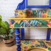 A vibrant, ornately decorated Vanity from the Vintage Victorian Mahogany Duchess Dressing Table Painted Bedroom Set sits against a white brick wall. The blue and yellow surfaces of the dressing table feature tropical-themed illustrations of toucans, foliage, monkeys, and various colorful plants. A potted plant rests on the floor to the left of this charming vanity.