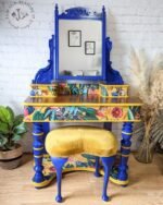 The Vintage Victorian Mahogany Duchess Dressing Table Painted Bedroom Set includes a vanity adorned with a large mirror featuring intricate blue trim. The wooden desk base is painted gold and boasts a vibrant floral design, complemented by matching blue legs and paired with a mustard yellow upholstered stool. Potted plants and framed wall art add decorative touches to the space.