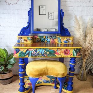 The Vintage Victorian Mahogany Duchess Dressing Table Painted Bedroom Set includes a vanity adorned with a large mirror featuring intricate blue trim. The wooden desk base is painted gold and boasts a vibrant floral design, complemented by matching blue legs and paired with a mustard yellow upholstered stool. Potted plants and framed wall art add decorative touches to the space.