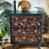 A dark blue chest of drawers with intricate red and orange paisley patterns sits against a white brick wall. It is adorned with decorative items, including a gold plate, a candle, and small sculptures. Surrounding the chest are potted plants and a lantern.