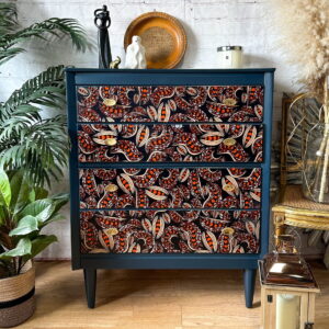 A wooden dresser with intricately patterned drawers featuring red, orange, and white designs against a dark background. The dresser sits on a wooden floor, surrounded by decorative plants, a wicker chair, a silver lantern, and various ornaments on top.