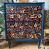 A vibrant navy blue chest of drawers with an intricate, colorful paisley design on the drawer fronts in shades of red, orange, and white. The surroundings include plants and various decor items, creating a stylish and lively atmosphere.