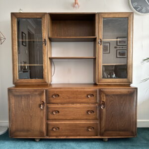 The Ercol Windsor Sideboard and Display Cabinets with Lighting in Golden Dawn Finish stands on a blue carpet. It features glass-paneled doors on the upper sections and solid doors on the lower sections, along with three central drawers and shelves in the middle. To the right, a wall clock is visible, and framed pictures adorn the wall.