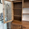 The Ercol Windsor Sideboard and Display Cabinets with Lighting in Golden Dawn Finish features a glass door, revealing empty shelves inside. Below the shelving area, the cabinet includes drawers and a wooden-door storage compartment. Positioned against a wall in a room, it stands next to a radiator cover and a standing lamp.