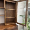 The Ercol Windsor Sideboard and Display Cabinet with Lighting in Golden Dawn Finish showcases two empty shelves behind an open glass door. The cabinet, featuring a polished finish, is set against a wall adorned with a clock, plant, and glass lantern in the backdrop.