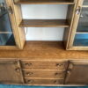 The Ercol Windsor Sideboard and Display Cabinets with Lighting in Golden Dawn Finish is showcased. It features a central section with three drawers beneath two open shelves, flanked by glass-fronted display sections on either side. The cabinet is set against a white wall, and the floor is covered with blue carpet.