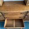 An Ercol Windsor Sideboard and Display Cabinet with a Golden Dawn finish, featuring multiple drawers and distinctive circular handles, stands on a blue carpeted floor. One drawer is open, revealing its empty interior divided by a single central divider.