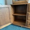 The Ercol Windsor Sideboard and Display Cabinet with Lighting in Golden Dawn Finish features a wooden structure with an open door, revealing two interior shelves. The cabinet is mounted with a mirror on top and is placed on a blue carpeted floor. Drawer handles are visible on the right side of the cabinet, and the interior appears empty.