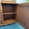 An Ercol Windsor Sideboard and Display Cabinet with Lighting, Golden Dawn Finish, with an open door revealing an empty interior shelf. The cabinet is situated on a blue carpet, and a green potted plant is partially visible in the background. The cabinet features a dark brown finish with natural wood grain visible.