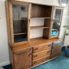 The Ercol Windsor Sideboard And Display Cabinets With Lighting in Golden Dawn Finish features glass doors and shelves on both sides, open compartments in the middle, and drawers and cabinets below. The sideboard stands on a teal carpeted floor with a wall clock visible in the background.