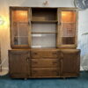 The Ercol Windsor Sideboard and Display Cabinets with Lighting in Golden Dawn Finish feature a mid-century modern design, combining open shelves and glass-fronted cabinets on the top with drawers and closed cabinets on the bottom. It is placed on a teal carpet, with a floor lamp and a wall clock visible in the background.