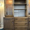 The Ercol Windsor Sideboard and Display Cabinets with Lighting in Golden Dawn Finish stand against the wall, showcasing glass-paneled doors on the upper left and right sides, with shelves centered in between. Below these, there are drawers and additional cabinet space. A floor lamp with a geometric design is positioned to the left. The floor features a blue carpet.