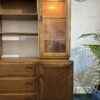 The Ercol Windsor Sideboard And Display Cabinets With Lighting in a Golden Dawn finish features a combination of drawers and shelves. On the right side, the upper section has a glass door with an illuminated interior, while the lower section has a wooden door. A wall clock is mounted above it, and a potted plant is partially visible on the right.