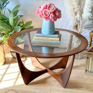 The G-Plan Astro Round Coffee Table Mid-Century Vintage, featuring a glass top and stylish wooden base, is set against a backdrop of various plants, pampas grass, and a white brick wall. On the table sits a blue vase filled with pink flowers and a stack of books. Sunlight filters through, creating a cozy atmosphere.