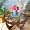 A G-Plan Astro Round Coffee Table Mid-Century Vintage with a wooden base stands in a well-lit room. On top of the table is a blue vase filled with pink flowers and two books. Surrounding the table are potted plants, a wicker chair, and decorative pampas grass.