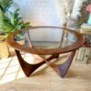 A G-Plan Astro Round Coffee Table Mid-Century Vintage with a sculpted wooden base sits on a hardwood floor. The background features a potted green plant, decorative pampas grass, and a wicker lamp beside a stack of books. The logo "Flip It & Restore It" is visible at the bottom left.