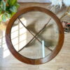 An overhead view of a G-Plan Astro Round Coffee Table Mid-Century Vintage on a wooden floor. The table features a circular wooden frame and cross-shaped wooden supports underneath the glass top. Beside the table, there is a potted plant with large green leaves, and a fluffy rug is nearby.