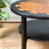 Here is a close-up of the MCM Circular Round 'Cog' Coffee Table by Nathan Furniture. This two-tier wooden side table features a top surface with a stained wood inlay showcasing geometric shapes, while the table legs and bottom tier are hand-painted in black. A green plant and light-colored flooring can be seen in the background.