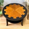 The MCM Circular Round 'Cog' Coffee Table by Nathan Furniture is a stunning wooden side table with a unique two-tone design. The tabletop features an orange-brown center with black, gear-like edges, while the base is painted black and includes a lower shelf. It sits beautifully on a wooden floor, surrounded by plants and decorative items.