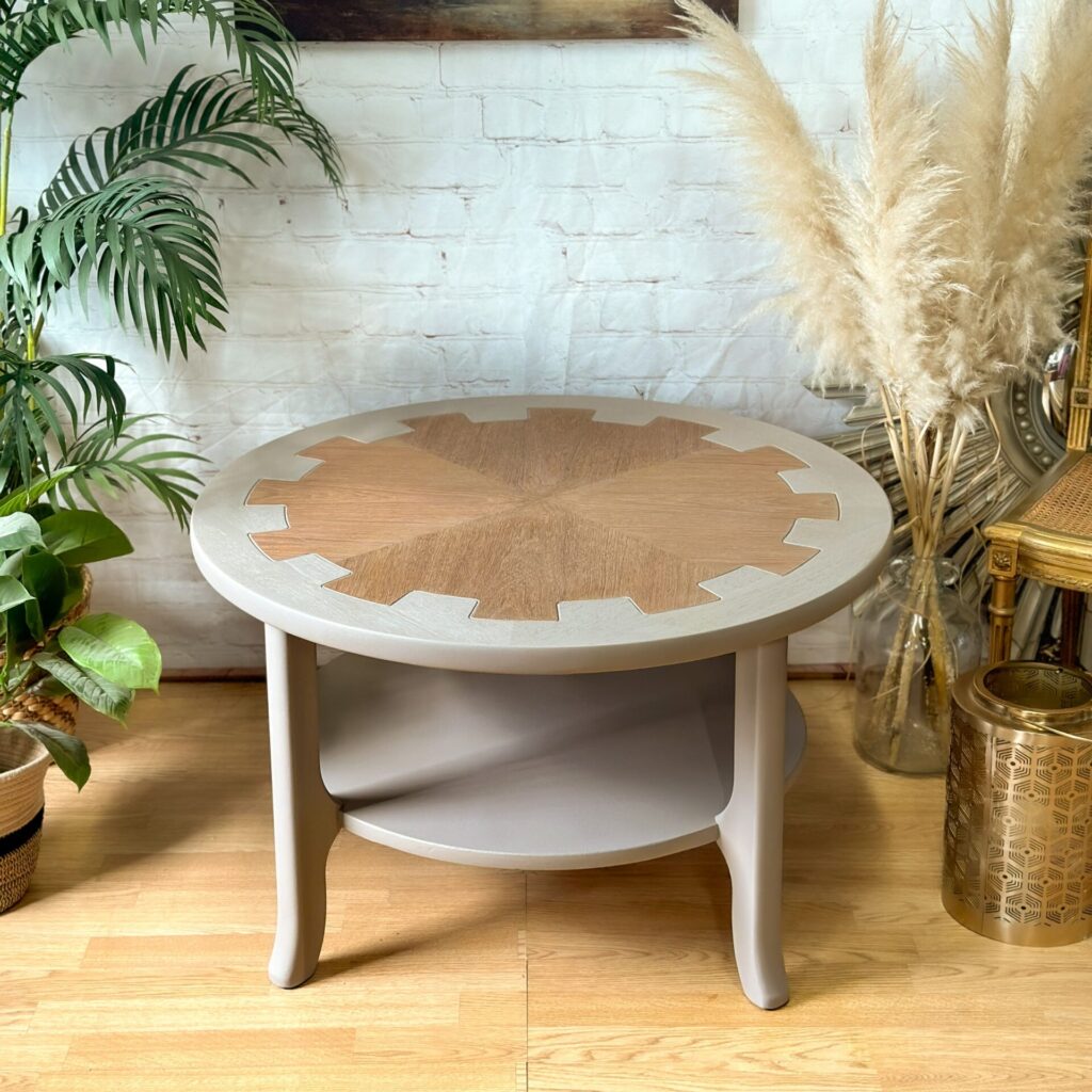 A MCM Circular Round 'Cog' Coffee Table by Nathan Furniture, hand-painted in soft khaki beige, stands on a light wood floor. It features a unique inlay pattern and a lower shelf. Surrounding the table are various green and dried plants, and a wicker basket, against a backdrop of white brick. A gold-toned accent piece is also nearby.