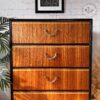 An Exquisite Mid Century MCM Walnut Tallboy with 6 drawers by Meredew, featuring a black frame and metal handles. The chest boasts drawers that showcase a warm, rich wood grain pattern. Above it, a framed abstract art print adorns the brick wall, while a tall houseplant is partially visible on the left side.