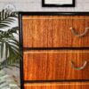 An Exquisite Mid Century MCM Walnut Tallboy 6 Drawers Chest Of Drawers by Meredew with a vertical grain pattern is seen against a white brick wall. It features six drawers with metallic handles. A potted plant with long, green leaves is partly visible on the left side, and a framed Henri Matisse print hangs on the wall above.