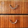 Close-up of two metallic, arch-shaped handles on the Exquisite Mid Century MCM Walnut Tallboy 6 Drawers Chest Of Drawers by Meredew, showcasing its rich brown wood grain. The handles are secured with small round mounts. In the top left corner, a faint circular logo with barely discernible markings can be seen.