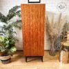 The Exquisite Mid Century MCM Walnut Tallboy 6 Drawers Chest Of Drawers by Meredew stands on black legs against a white brick wall. It is flanked by green plants on the left and dried pampas grass in a vase on the right. A small, mirrored stool is partially visible on the right side.