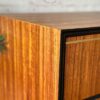 Close-up of the Exquisite Mid Century MCM Walnut Tallboy 6 Drawers Chest Of Drawers by Meredew, showcasing its smooth, lacquered finish. The corner of the chest is prominently featured, highlighting its precise edges and rich wood grain. Above the furniture is a circular logo with text encircling a paintbrush and hammer.