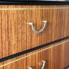 Close-up of an "Exquisite Mid Century MCM Walnut Tallboy 6 Drawers Chest Of Drawers by Meredew" featuring six drawers, each adorned with metallic handles. The wood grain runs horizontally, accented by thin black and gold inlays between the drawers. The top edge is finished in black, and the background displays a segment of a "Flip It & Restore It" logo.