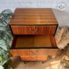 The Exquisite Mid Century MCM Walnut Tallboy 6 Drawers Chest of Drawers by Meredew stands against a white brick wall. The top drawer is closed while the middle drawer is open, revealing an empty interior. A potted plant is situated to the left and a wicker decorative piece is on the right side.