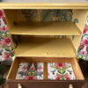 A hand-painted tropical-themed cabinet featuring open yellow shelves and a brown drawer at the bottom, with the inside back and drawer adorned in wallpaper showcasing large leaves, pink flowers, and elephants. The background of the image is also decorated with floral patterns.