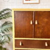 The Hand-painted Tropical Tallboy stands against a white brick wall, featuring a warm brown and yellow color scheme. It has two upper doors and three lower drawers, all adorned with leather pulls. To the left of the cabinet is a large potted plant, and above it hangs a framed painting of a Highland cow.