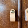 Close-up of a hand-painted, upcycled wooden cabinet featuring a light tan leather pull secured by a round, gold-colored knob. The leather pull is positioned vertically on the cupboard door. The wood has a rich, dark brown finish with visible grain texture.