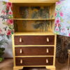 A refurbished yellow "Hand-painted cabinet, Tallboy, Linen Cupboard, Upcycled Cupboard" with three brown drawers featuring leather handles. The cabinet's open doors reveal tropical-patterned wallpaper inside with plants and a few animals. Part of a patterned wall can be seen in the background, along with a potted plant. Perfect for bedroom storage or kitchen storage.