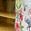 Close-up of a hand-painted Tallboy cabinet featuring colorful, detailed illustrations of tropical wildlife including a leopard, hummingbird, and elephant among vibrant flowers and greenery. The "Flip It & Restore It" logo with a hammer and paintbrush is visible in the corner.