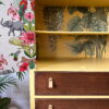 The Hand-painted Tallboy Linen Cupboard presents a vibrant, jungle-themed design. The left side showcases lively wallpaper adorned with a parrot, an elephant, and flowers. The open cabinet reveals shelves decorated with tropical leaves. Below, three wooden drawers feature light brown handles, adding to its unique charm.