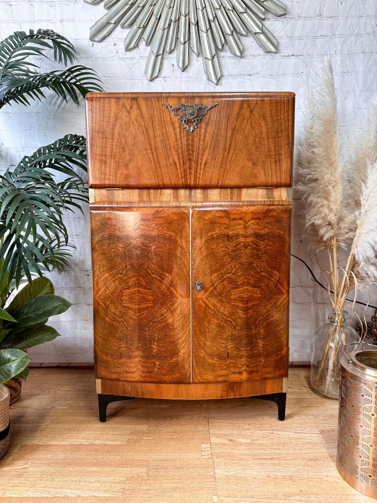 A Rivington Cocktail Cabinet, a vintage drinks bar from the 1950s with a stunning green and gold Art Deco design and featuring walnut grain adorned with monstera leaf patterns, stands against a white brick wall. The retro bar boasts two lower doors and a smaller upper compartment with a decorative emblem. Surrounding the cabinet are lush plants and ornamental grasses.