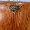 A close-up of the Rivington Cocktail Cabinet, an ornate wooden piece from the 1950s with a vintage art deco aesthetic. The cabinet, made from walnut and featuring green and gold accents, showcases a decorative metal embellishment in the center with intricate scrolling patterns and a floral design. Above, part of a sunburst mirror accented by metal rays is visible.