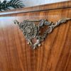 Close-up of an ornate, brass corner embellishment on a wooden piece of furniture. The intricate design features floral and scroll elements. A green leafy Monstera plant is partially visible in the background, and there are white brick wall details behind the Rivington Cocktail Cabinet, a vintage drinks bar with an art deco style from the 1950s, finished in green and gold walnut.